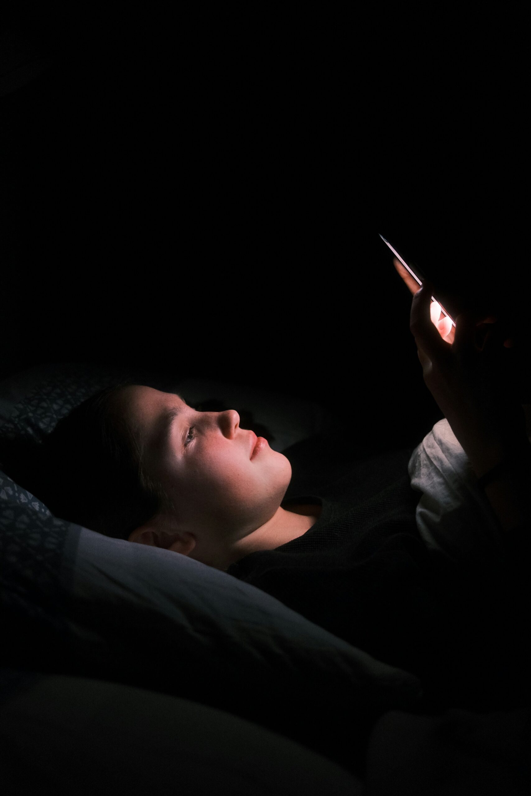 kajetan-sumila-ygB6yVjb7a4-unsplash a person using their phone in bed at night. the night is shining on their face while the rest of the room is dark